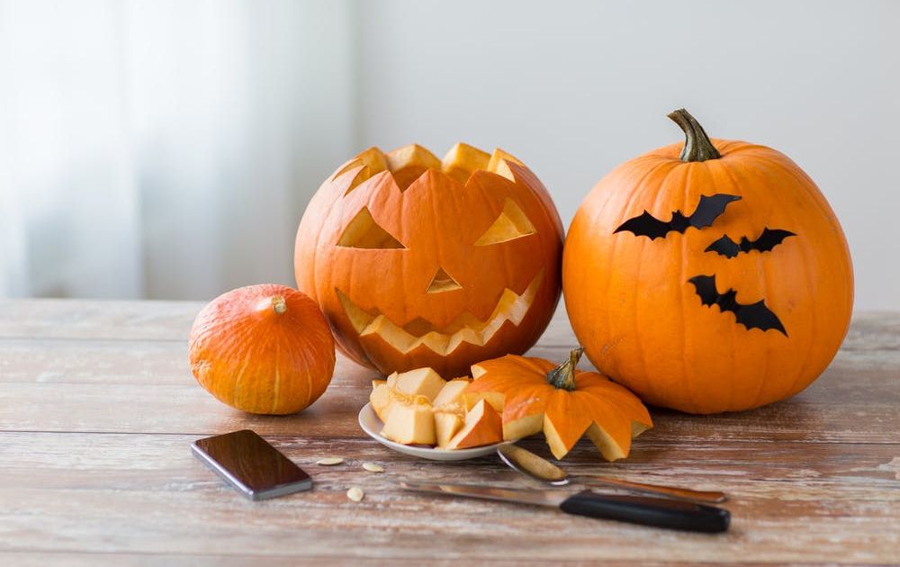 Een close-up van een houten tafel vol Halloween-versieringen, popcorn in een spinnenweb-schaal en kleine pompoenemmers, met mensen in kostuums vaag op de achtergrond.