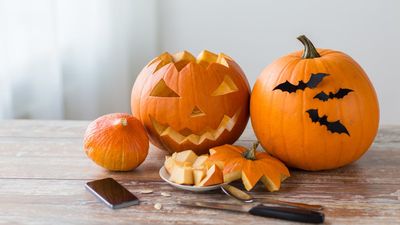 Een close-up van een houten tafel vol Halloween-versieringen, popcorn in een spinnenweb-schaal en kleine pompoenemmers, met mensen in kostuums vaag op de achtergrond.
