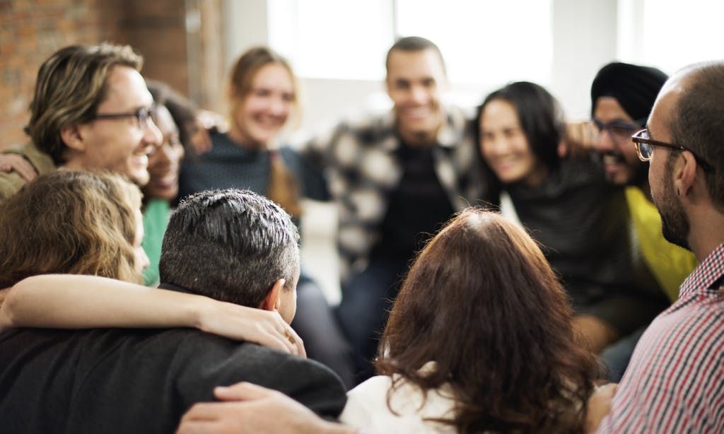 A diverse group of friends or colleagues sitting closely in a circle with their arms around each other's shoulders, smiling and laughing together.