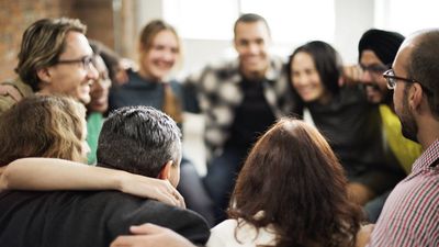 A diverse group of friends or colleagues sitting closely in a circle with their arms around each other's shoulders, smiling and laughing together.