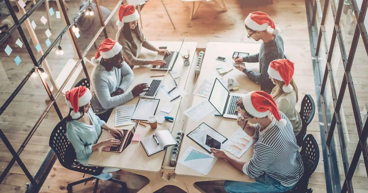 A high-angle view of six diverse colleagues working at a long wooden desk in a modern office, all wearing red and white Santa hats during the holiday season.
