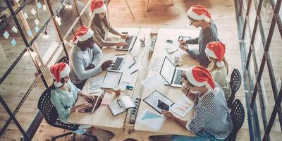 A high-angle view of six diverse colleagues working at a long wooden desk in a modern office, all wearing red and white Santa hats during the holiday season.