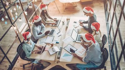 A high-angle view of six diverse colleagues working at a long wooden desk in a modern office, all wearing red and white Santa hats during the holiday season.