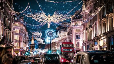 A night view of Regent Street in London during the holiday season, illuminated by large, glowing angel Christmas lights spanning across the busy road filled with black cabs and a red double-decker bus.