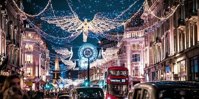 A night view of Regent Street in London during the holiday season, illuminated by large, glowing angel Christmas lights spanning across the busy road filled with black cabs and a red double-decker bus.