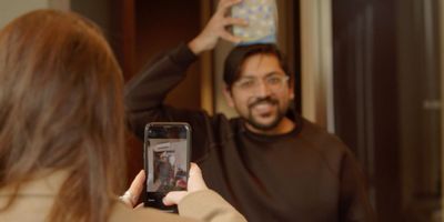 A colleague photographing a smiling teammate on a phone as he balances a cushion on his head, taking part in an indoor office team building challenge.