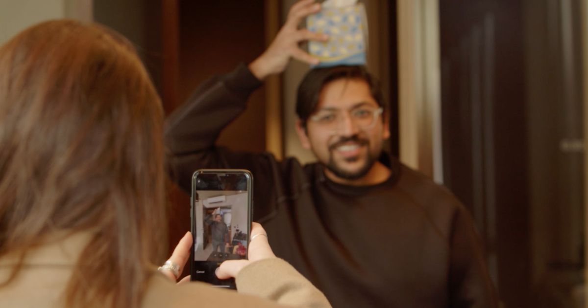 A colleague photographing a smiling teammate on a phone as he balances a cushion on his head, taking part in an indoor office team building challenge.