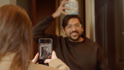 A colleague photographing a smiling teammate on a phone as he balances a cushion on his head, taking part in an indoor office team building challenge.