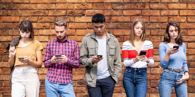 Een diverse groep van vijf jonge volwassenen die naast elkaar staan voor een getextureerde bakstenen muur, elk aandachtig kijkend naar het scherm van hun smartphone.