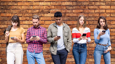 Een diverse groep van vijf jonge volwassenen die naast elkaar staan voor een getextureerde bakstenen muur, elk aandachtig kijkend naar het scherm van hun smartphone.
