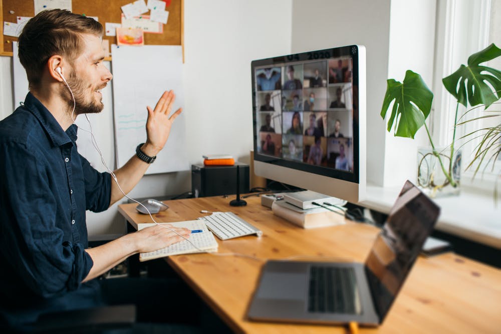 Vue de profil d'un homme portant des écouteurs, saluant son écran d'ordinateur de bureau tout en animant ou en participant à une grande réunion vidéo de groupe depuis son bureau à domicile.