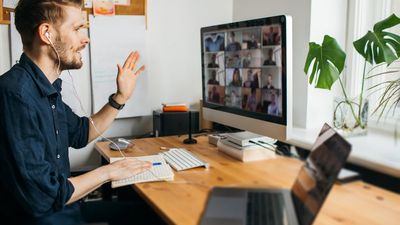 Een zijaanzicht van een man met een koptelefoon op, die zwaait naar zijn desktopmonitor terwijl hij een grote groepsvideovergadering host of eraan deelneemt vanuit zijn thuiskantoor.