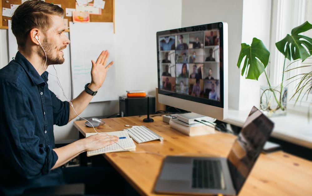 Een zijaanzicht van een man met een koptelefoon op, die zwaait naar zijn desktopmonitor terwijl hij een grote groepsvideovergadering host of eraan deelneemt vanuit zijn thuiskantoor.