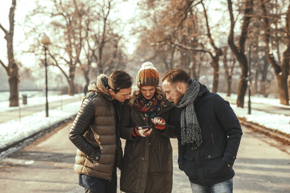 Trois amis en manteaux d'hiver chauds et bonnets, debout ensemble sur un chemin dans un parc, regardant attentivement un smartphone tenu par une femme au centre.