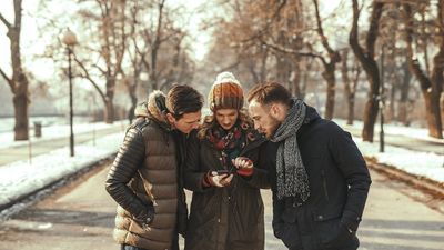 Drie vrienden in warme winterjassen en mutsen, samen staand op een pad in een park, aandachtig kijkend naar een smartphone die een vrouw in het midden vasthoudt.