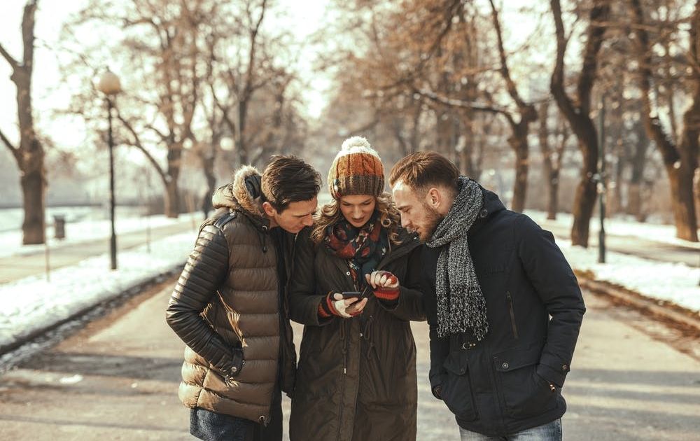 Drie vrienden in warme winterjassen en mutsen, samen staand op een pad in een park, aandachtig kijkend naar een smartphone die een vrouw in het midden vasthoudt.