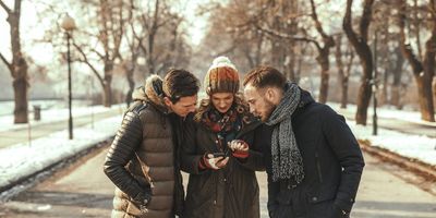 Trois amis en manteaux d'hiver chauds et bonnets, debout ensemble sur un chemin dans un parc, regardant attentivement un smartphone tenu par une femme au centre.