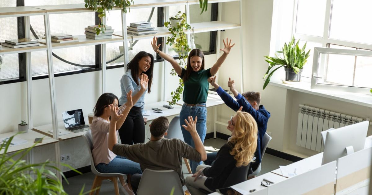Bovenaanzicht van een creatief team van zes collega's die een succes vieren in een licht kantoor, met opgeheven handen, glimlachend en klappend samen.