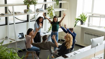 Bovenaanzicht van een creatief team van zes collega's die een succes vieren in een licht kantoor, met opgeheven handen, glimlachend en klappend samen.
