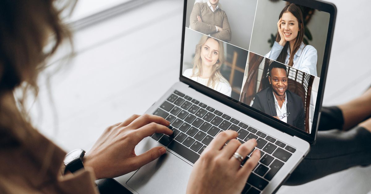 An over-the-shoulder view of a woman typing on her laptop while participating in a virtual meeting with four smiling colleagues displayed in a grid on her screen.