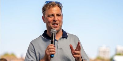 A Wildgoose host speaking into a handheld microphone outdoors under a clear blue sky, sunglasses resting on his head, addressing a group at an event.