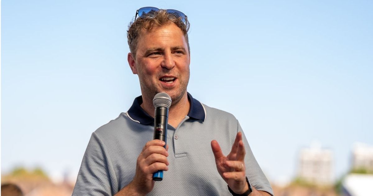 A Wildgoose host speaking into a handheld microphone outdoors under a clear blue sky, sunglasses resting on his head, addressing a group at an event.