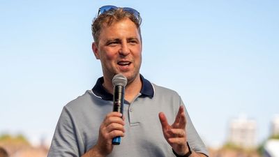 A Wildgoose host speaking into a handheld microphone outdoors under a clear blue sky, sunglasses resting on his head, addressing a group at an event.