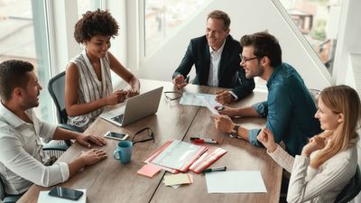 Bovenaanzicht van een divers team van vijf glimlachende professionals die samenwerken rond een houten vergadertafel in een licht en modern kantoor.