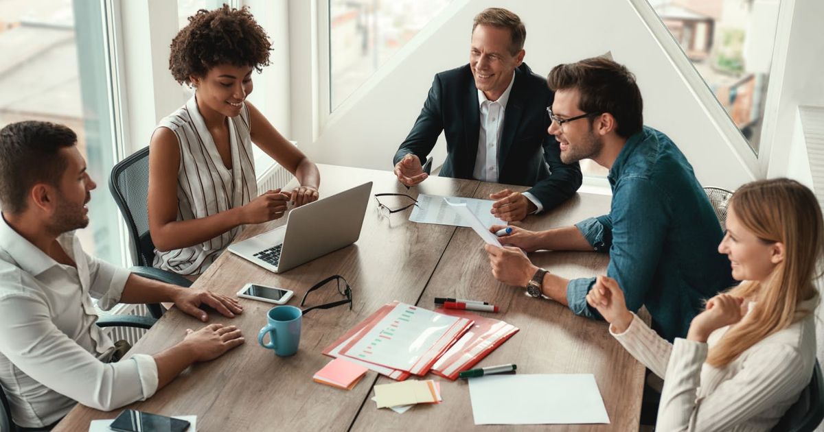 Bovenaanzicht van een divers team van vijf glimlachende professionals die samenwerken rond een houten vergadertafel in een licht en modern kantoor.