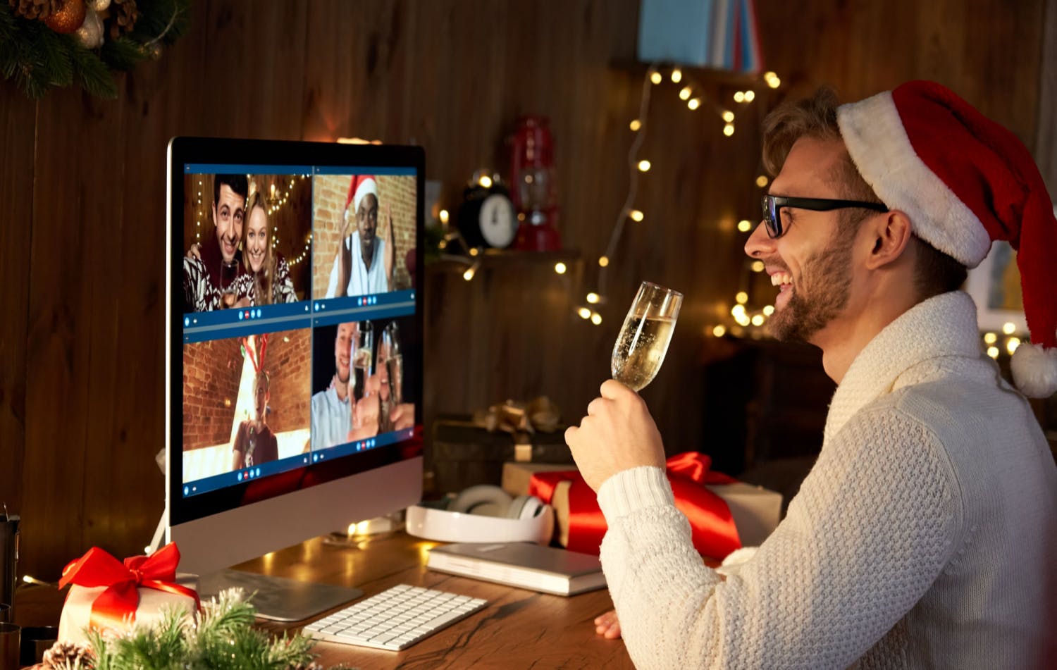 Un homme souriant coiffé d'un bonnet de Père Noël est assis à son bureau et lève un verre de champagne pour porter un toast à ses collègues lors d'une célébration de Noël virtuelle sur l'écran de son ordinateur.