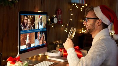 Een glimlachende man met een kerstmuts zit aan zijn bureau en heft een glas champagne om zijn vrienden te toasten tijdens een virtuele kerstviering op zijn computerscherm.