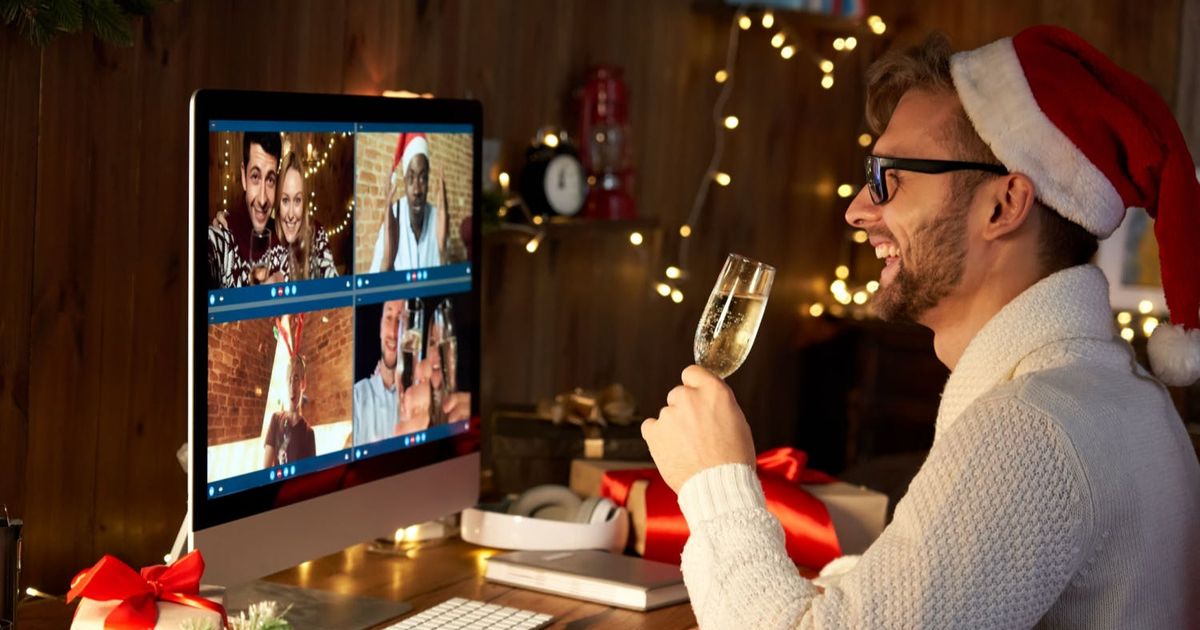 Een glimlachende man met een kerstmuts zit aan zijn bureau en heft een glas champagne om zijn vrienden te toasten tijdens een virtuele kerstviering op zijn computerscherm.