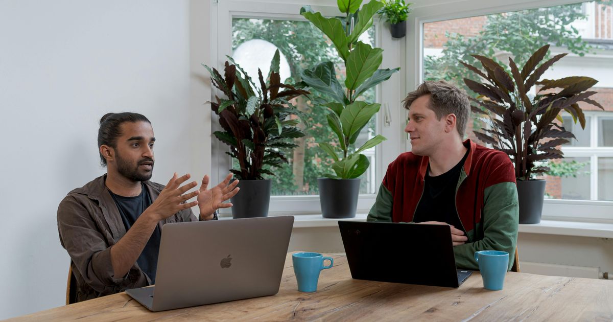 Two men sitting at a wooden table with laptops and blue mugs, having an engaged conversation in a bright office filled with large potted plants.