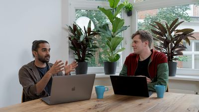 Two men sitting at a wooden table with laptops and blue mugs, having an engaged conversation in a bright office filled with large potted plants.