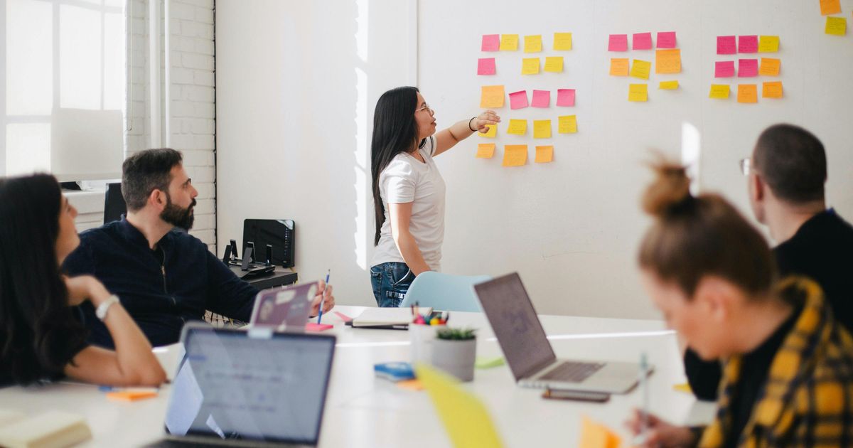 A businesswoman leading an office brainstorming session, pointing to organized rows of colorful sticky notes on a white wall while colleagues watch from a conference table.