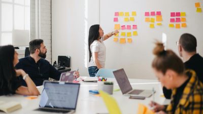 A businesswoman leading an office brainstorming session, pointing to organized rows of colorful sticky notes on a white wall while colleagues watch from a conference table.