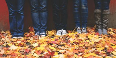 Four people standing side by side against a red wall in jeans, trainers and boots, photographed from the knees down on a pavement thickly covered with yellow and orange autumn leaves.