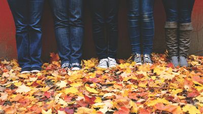 Four people standing side by side against a red wall in jeans, trainers and boots, photographed from the knees down on a pavement thickly covered with yellow and orange autumn leaves.